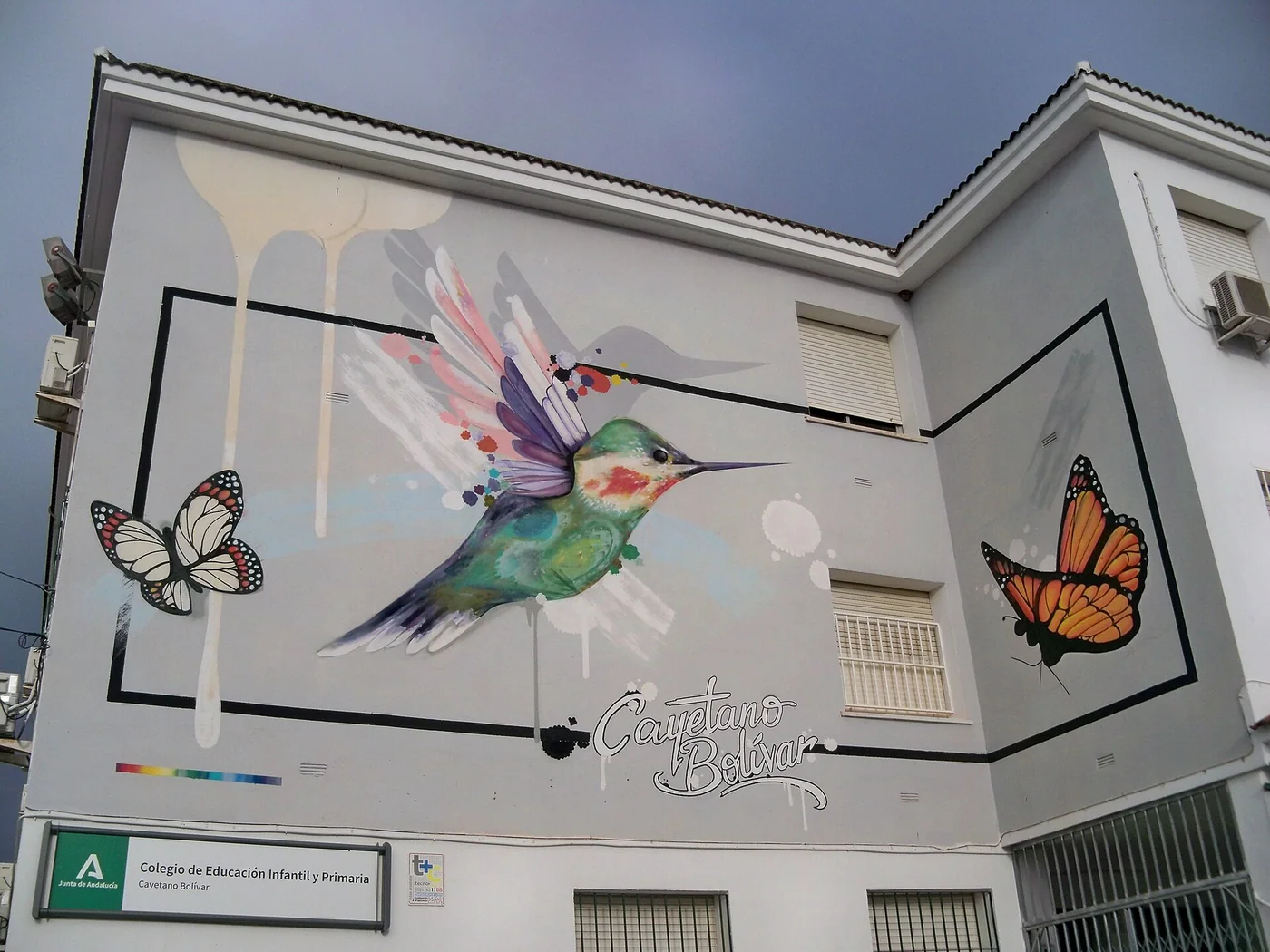 Mural de Rubén de marzo de 2022 en el colegio de educación infantil y primaria Cayetano Bolívar, en el número 2 de la calle del Romero, barrio Santa Rosalía, Málaga, España. El mural contiene el nombre del colegio Cayetano Bolívar junto a la imagen de un colibrí y dos mariposas. [1]