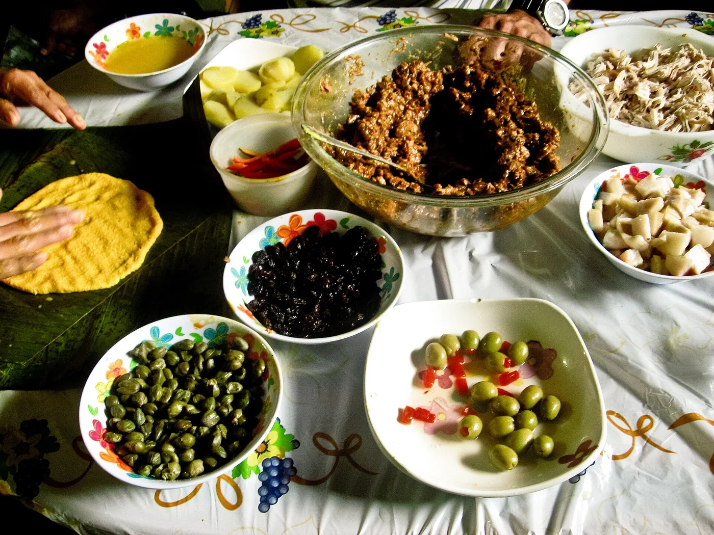 Woman spreading maize dough on a banana leaf while preparing Venezuelan hallacas, beside bowls of meat stew, raisins, olives, capers, chopped ingredients, and additional dough for Christmas.