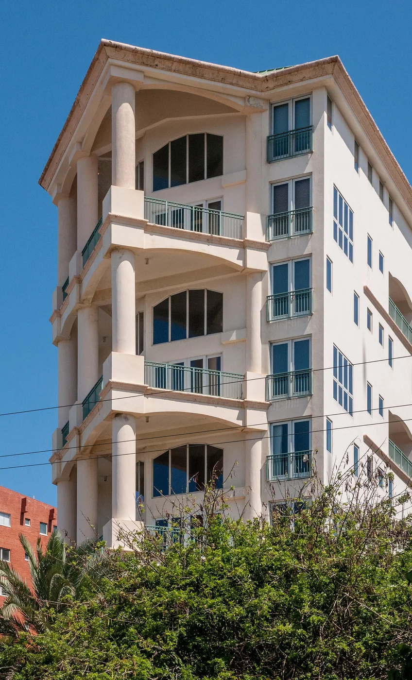 White multi-story apartment building in El Morro, Porlamar, Margarita Island, Venezuela, with deep stacked balconies framed by broad arches and round columns, green metal railings, corner windows, and a peaked roofline.